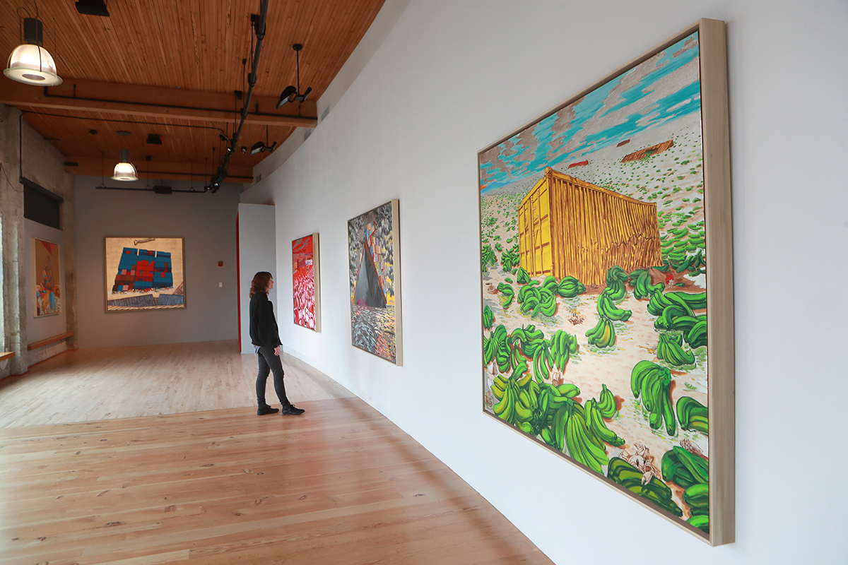 Image of large, brightly colored paintings in an art gallery with wooden floor and person in black coat viewing the work. 