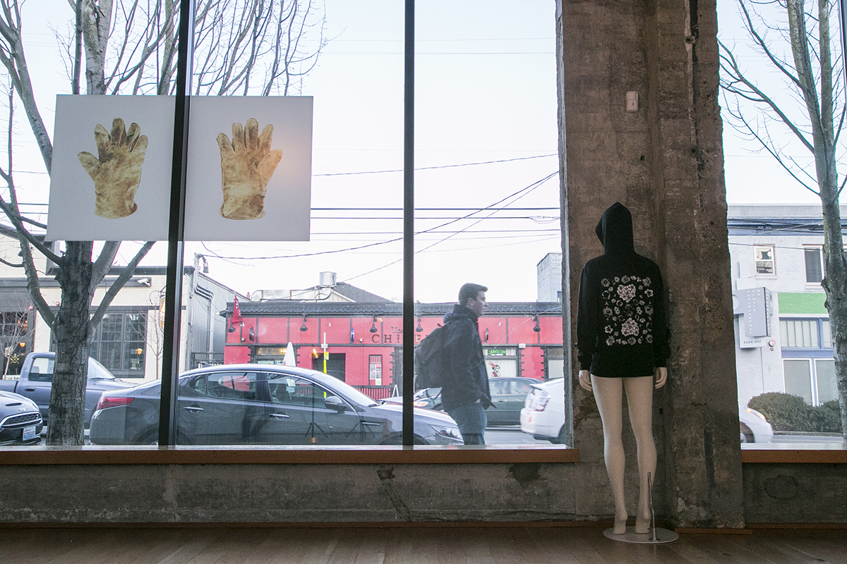 Two artworks in front of windows, on left image of yellow gloves, on right, mannequin with black hoodie and floral print