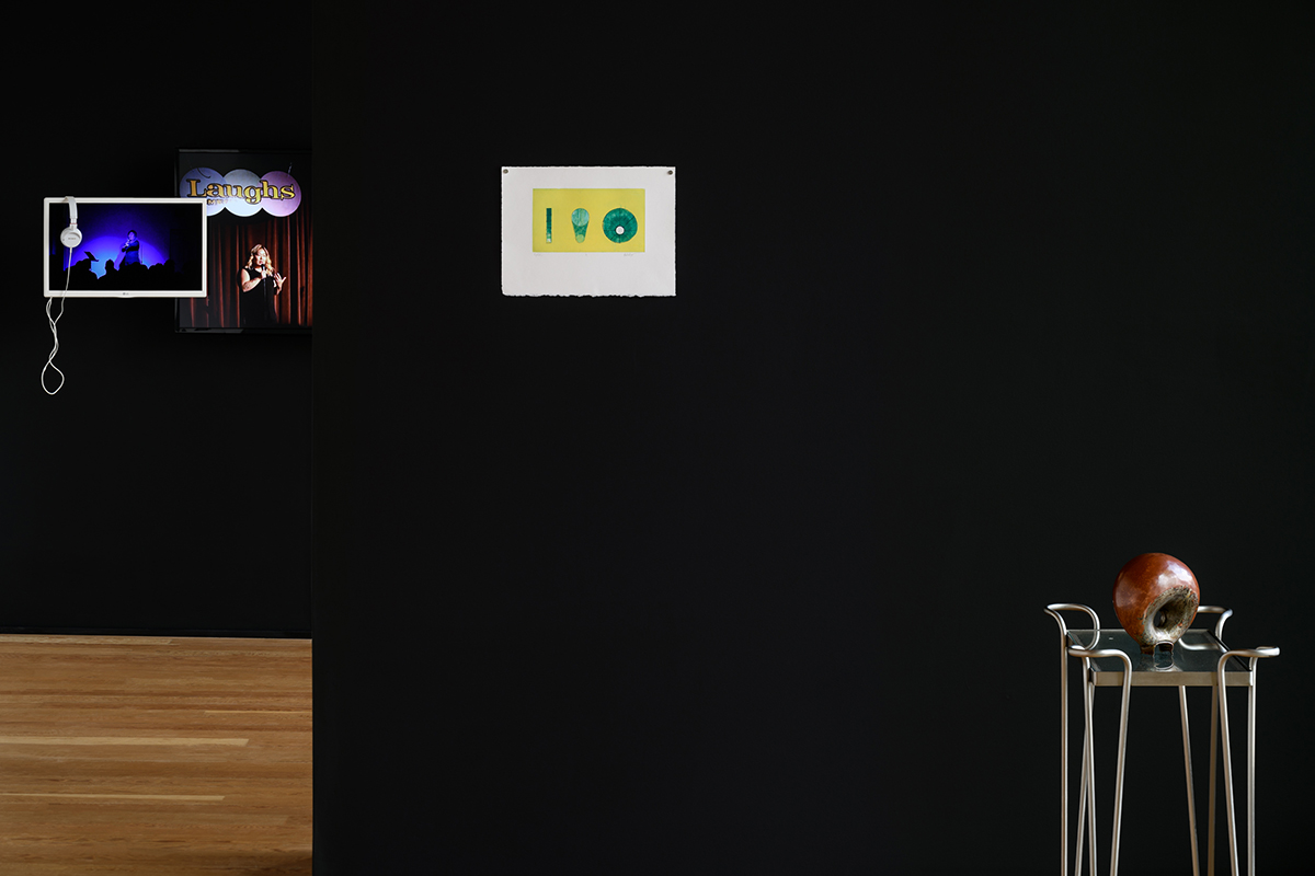 Three artworks on black walls. Comedy on video monitors, yellow and green etching, and copper, glass, steel sculpture