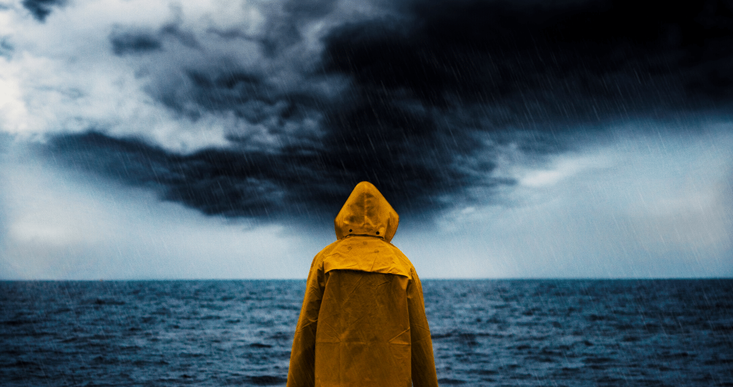 A person in a yellow rain jacket stands in the rain, looking out over a storm-clouded sky above a body of water.