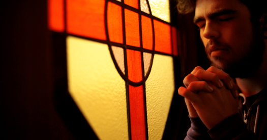A person with short brown hair and a bead folds hands in prayer beside a yellow and red stained glass windwo.