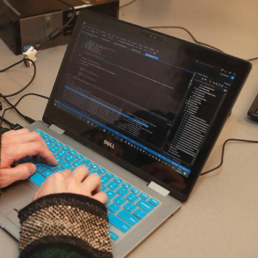 hands on a blue laptop keyboard with a dark screen with lines of code on a baige desk.