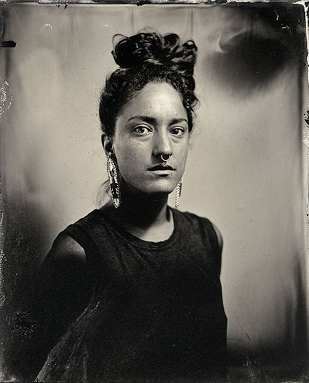 a black and white image of the artist. The artist is facing the camera, wearing earrings and has long hair up on their head.