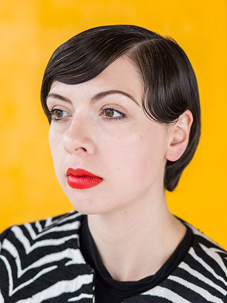 a portrait of a woman with light skin, short dark hair, red lips, in a striped shirt on a yellow background