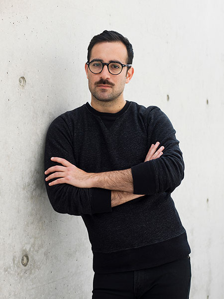 A photograph of artist Rafael Soldi, leaning against a wall with arms crossed over chest, wearing a dark sweater and glasses. 
