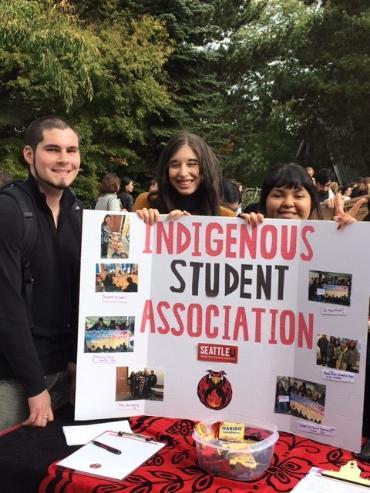 Three native students standing behind a sign on a table