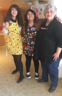 Two students, Macheddie and Hanna-Marie, standing with elder Jeanne Raymond wearing aprons from cooking the community dinner