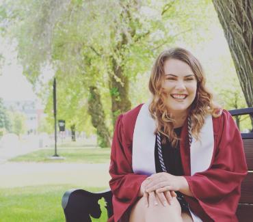 Student in a red graduation gown and white stole sitting on a bench
