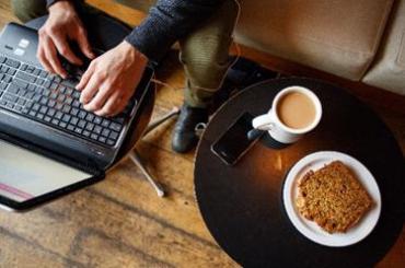 Arial view of small table with coffee and pastry and a person sitting on a couch with their computer on another table