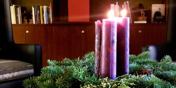 A wreath three lit purple candles in the middle and a bookshelf in the background