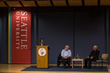 Lucas Sharma, SJ at the podium while, Greg Boyle, SJ, and Pat Howell, SJ, sit on the stage
