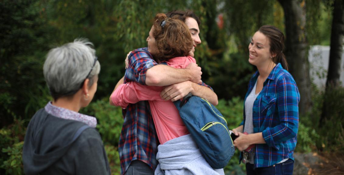 A student giving a person a hug while another person smiles in background. 