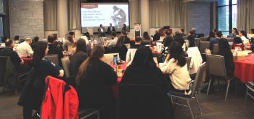 A conference with the audience around tables listening to three people on stage.