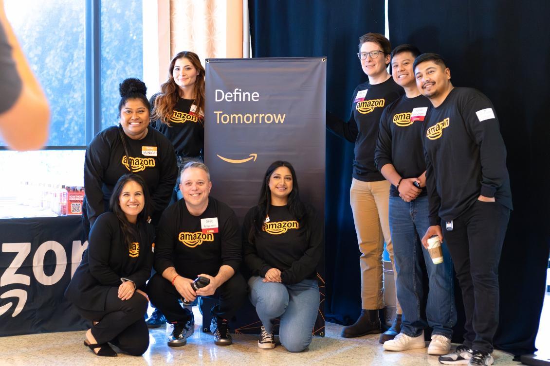 Amazon representatives and alumni connect with students at the Albers Business and Engineering Career Fair, posing together beside their booth’s Define Tomorrow display.