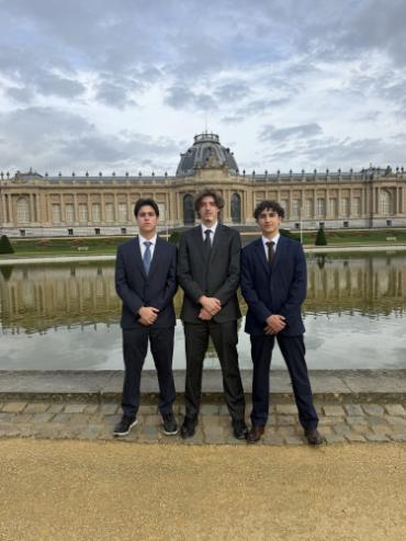 Lars Kaser and two classmates in dark suits stand before a reflective pond outside the Royal Museum for Central Africa in Tervuren, Belgium.