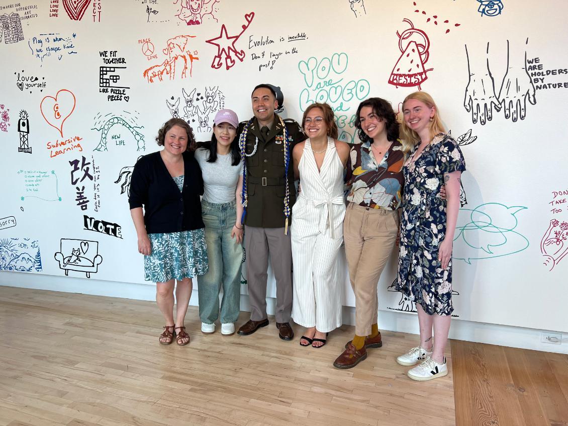 Six smiling students stand in front of wall with artistic writing