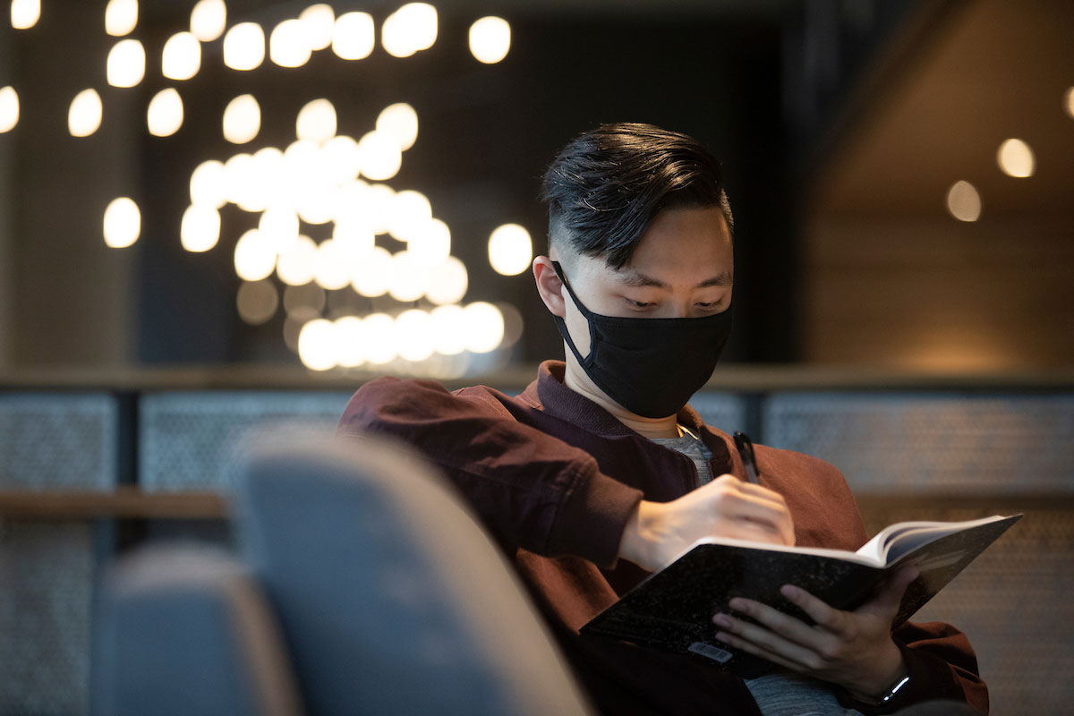 Student, wearing a mask, does some writing in a notebook in one of the residence halls.