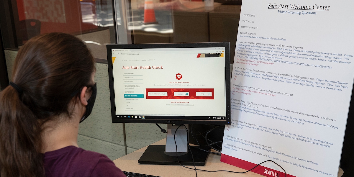 A student sits at computer in Safe Start Welcome Center with Safe Start Health Check web page on screen.