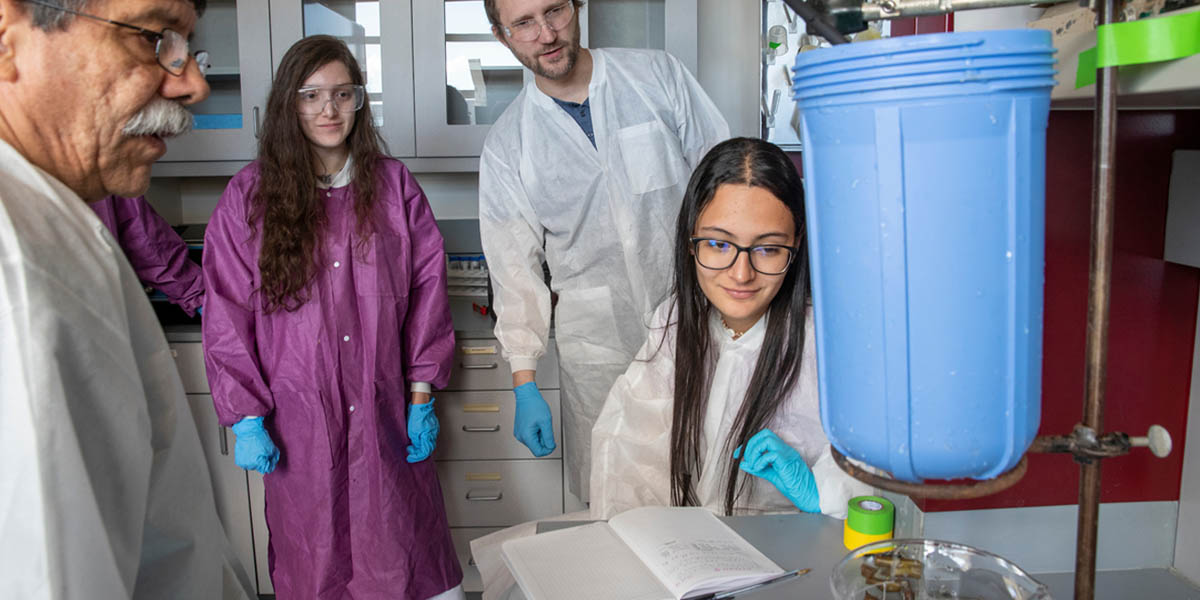 Two students and two faculty members wearing protective lab coats and eyewear while reviewing research results in a lab.