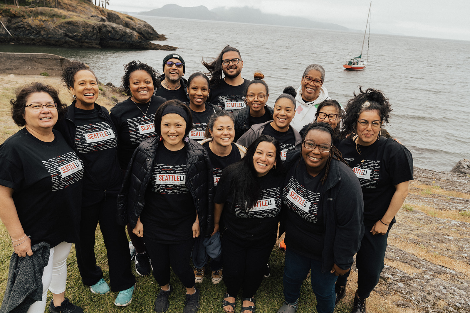 15 BIPOC individuals standing on a beach, smiling and all wearing black Seattle U t-shirts. The sky is grey. 