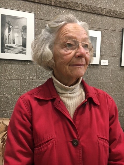 Gray haired woman (Susanna Hayes) with a white turtleneck sweater under a red collared jacket