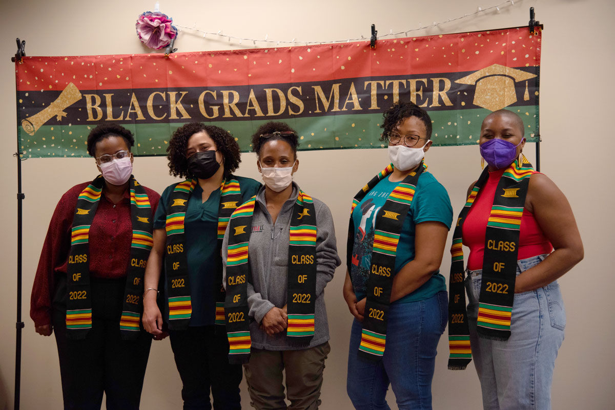 A group of black graduates wearing special Class of 2022 academic stoles pose under a Black Grads Matter banner.