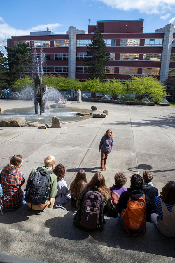 A class of students sitting on steps facing a professor standing in front of a bronze fountain and trees on a sunny day