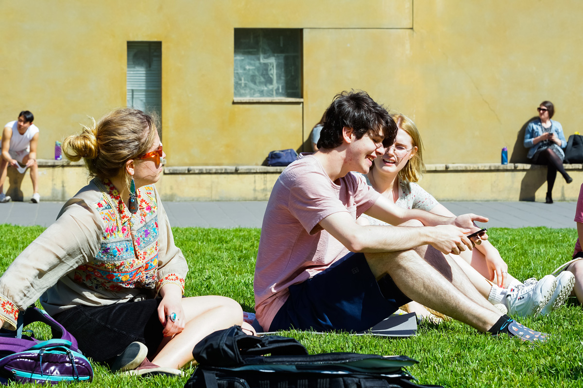 A group of students wearing shorts and sneakers soak up some sun on a green lawn and others sit against the chapel wall